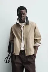 Sobrecamisa de corte relajado color beige, con cuello solapa en efecto piel de color negro. Presenta cierre frontal con cremallera, bolsillos de vivo en la cadera y mangas largas con puños ajustables.