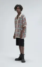 Camisa de manga corta, tejida a crochet con un patrón de cuadrados en colores beige, naranja y turquesa. Presenta cuello tipo polo y cierre frontal con botones.