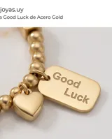 Pulsera elástica de acero dorado compuesta por pequeñas esferas, con un dije en forma de corazón y una placa rectangular con la inscripción 'Good Luck'.