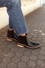 Botas tipo Chelsea de cuero negro, con paneles laterales elásticos, detalle de textura en el talón y suela plana con detalle en madera clara.