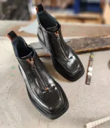 Bota corta de cuero negro con diseño de punta cuadrada y suela track. Presenta un cierre frontal central, detalles de pespuntes y una textura combinada en los laterales.