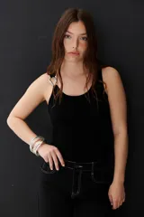 Musculosa negra ajustada al cuerpo con escote redondo y tirantes finos. Presenta un detalle metálico en forma de aro o hebilla en la unión del tirante con el cuerpo de la prenda.