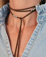 Collar tipo corbatín confeccionado en tiento de cuero marrón, con detalle de cuentas metálicas plateadas distribuidas a lo largo de las tiras.