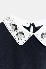 Jersey de punto negro de manga corta con cuello solapa blanco en contraste, decorado con abalorios joya y lentejuelas. Presenta cierre trasero con abertura y botón.