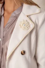 Blazer cruzado color beige, con cierre de cuatro botones frontales. Presenta detalles artesanales de encaje con perlas y cristales en la solapa y en los bolsillos delanteros.