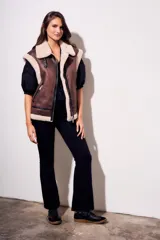 Chaleco oversize estilo gamulán en color marrón, confeccionado en cuero vegano con forro y solapas de corderito sintético en contraste. Posee cierre frontal metálico, bolsillos con cremallera y detalles de correas con hebillas en el cuello.