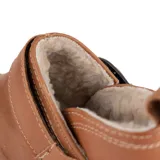 Botita baja Marcel confeccionada en cuero vacuno color marrón, con interior de corderito ultrasuave. Presenta costuras pespunteadas decorativas, tira lateral con hebilla metálica dorada y detalles de elástico negro. Suela plana de goma negra.