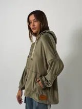 Canguro de plush color verde oliva, con capucha, cordones ajustables, bolsillo frontal tipo canguro y mangas con puños elásticos. Presenta un parche de cuero sintético con logo en la parte inferior.