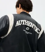 Campera bomber de cuero sintético con diseño bicolor, cuerpo negro y mangas blancas. Presenta cuello, puños y cintura con tejido elástico a rayas, cierre frontal con botones a presión y bordado con la palabra Authentic en el pecho.