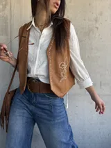 Chaleco de gamuza color camel con bordados en hilo crudo a lo largo de los bordes delanteros, diseño floral bordado en el lateral, tachas metálicas decorativas y botones a presión ocultos.