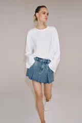 Falda corta de tiro medio confeccionada en tejido denim de algodón. Presenta diseño con tablas, bolsillos delanteros, cintura con trabillas y bajo desflecado. Cierre frontal con cremallera, botón y ganchos metálicos.