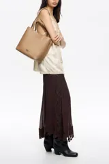 Bolso de hombro tipo tote bag de piel vacuna color beige, con doble asa de mano y correa ajustable de tejido. Presenta un cierre de cordón en la parte superior y un logo metálico en relieve en el frente. Incluye bolsillo interior con cierre de cremallera.