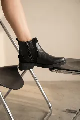 Bota corta de cuero negro con taco cuadrado bajo. Presenta un diseño versátil con un accesorio desmontable que incluye flecos de cuero, tachas metálicas y una cadena decorativa alrededor del tobillo.