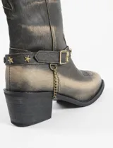Botas estilo tejano de cuero vegano en tono marrón con efecto desgastado. Presentan costuras decorativas, una correa ajustable con tachas en forma de estrellas y una pequeña cadena colgante en el lateral.