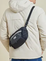 Bandolera negra con diseño técnico, confeccionada en poliamida con textura de cuadrícula. Cuenta con doble cierre frontal, bolsillos laterales de malla y correa ajustable. Incluye el logo de la marca Rusty en el frente.