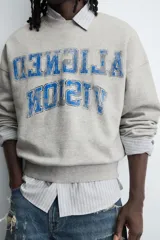 Sudadera gris de corte boxy fit, con cuello redondo y manga larga. Presenta un estampado frontal con texto en azul con efecto desgastado y acabados en rib en puños y ruedo.