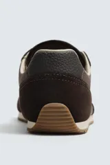Zapatilla deportiva estilo barefoot, color marrón, con diseño de puntera ancha y redondeada. Confeccionada en una combinación de materiales textiles y detalles en serraje. Cuenta con suela fina de goma flexible y cordones en color crema.