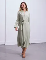 Vestido maxi color beige, con cuello mao y escote en V. Presenta mangas largas con presilla y botón para regular el largo, y un lazo ajustable en la cintura que marca la silueta.