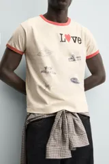 Camiseta de manga corta color crema con cuello redondo y ribetes en color rojo. Presenta un estampado frontal con la palabra "Love" donde la 'o' es un corazón rojo, y varios parches gráficos en tonos grises y negros.