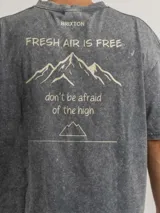 Camiseta de corte oversize con efecto lavado, color gris oscuro. Presenta un estampado gráfico en la espalda con el logo de la marca Brixton y la frase 'Fresh Air Is Free, don't be afraid of the high' junto a un diseño de montañas.
