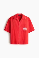 Camisa de manga corta color rojo con cuello abierto en contraste blanco, cierre frontal de botones y un bolsillo en el pecho con estampa gráfica.