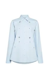 Camisa de algodón color celeste con cuello clásico, manga larga con puños y cierre frontal de botones. Presenta apliques decorativos de pedrería brillante distribuidos por toda la prenda.
