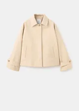 Chaqueta corta de algodón color beige, de corte holgado, con cuello clásico, cierre frontal de botones y bolsillos ribeteados. Cuenta con mangas largas amplias con puños ajustables mediante tiras y botones.