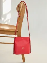 Cartera tipo bandolera de cuero color rojo, con diseño minimalista, estructura rectangular y correa larga ajustable con nudos. Presenta un logo metálico grabado en la parte frontal.