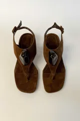 Sandalia de gamuza marrón con taco cuadrado bajo, diseño de tira tipo dedo y aplique decorativo negro brillante en el empeine. Cuenta con tira ajustable en el talón con hebilla.