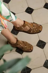 Sandalias guillermina de cuero gamuza en color camel, estilo cangrejera, con tiras cruzadas decoradas con tachas metálicas doradas y hebilla de ajuste en el tobillo.