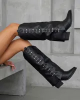 Botas texanas de caña alta en color negro, confeccionadas en cuero sintético. Presentan un diseño con detalle de ojales metálicos alineados verticalmente a lo largo de la caña, punta fina y taco cuadrado de altura media.