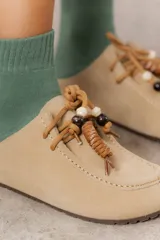 Zueco de cuero gamuzado color tostado, con cordones decorativos adornados con cuentas de madera y piezas envueltas en cuero. Diseño tipo mocasín con costuras visibles en la parte superior y abierto en la parte trasera.