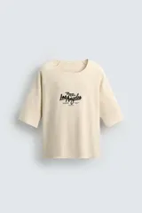 Camiseta de hombre, corte relaxed fit, color beige claro con textura. Presenta cuello redondo amplio y manga corta. Estampado frontal en negro con texto que incluye "Los Angeles".