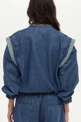 Camisa de denim azul con bordados blancos en el frente y las hombreras. Cuenta con cuello mao y cierre frontal con botones a presión.