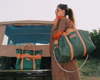 Bolso tipo Duffle o bolso deportivo de lona color verde militar, combinado con asas cortas, correa larga ajustable y detalles en cuero de color tostado. Cuenta con la marca grabada en un aplique frontal.