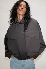 Chaqueta estilo bomber de color gris oscuro, con cuello subido, manga larga y corte holgado. Presenta bolsillos delanteros con solapa, bajo con pliegues y cierre frontal con cremallera oculta y botones a presión.