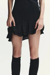 Vestido corto negro de tejido fluido con cuello halter que se ata con lazada. Tiene forro interior, detalle de frunces y bajo asimétrico con volantes. Cierre lateral con cremallera oculta.
