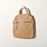 Mochila color beige con cierre superior y bolsillo frontal con cierre. Tiene doble asa corta y correas traseras regulables. Incluye un dije colgante de color rosa.