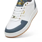 Championes Puma Court Classic de diseño urbano, color blanco con detalles en negro en la lengüeta y el talón. Presenta logo Puma en dorado en el lateral y la lengüeta, ojales superiores metálicos dorados y suela de goma color caramelo.