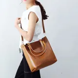 Bolso tipo tote bag de eco cuero color marrón caramelo, con dos asas de mano y correa larga ajustable para llevar al hombro o cruzado. Presenta un bolsillo exterior frontal.