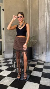 Falda tejida a mano en crochet, con diseño a rayas horizontales en tonos marrón y negro con hilos de lurex dorado. El ruedo presenta una cascada de flecos largos y finos en color negro.