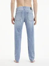 Jeans de mezclilla azul claro, tiro medio, con corte slim que se estrecha hacia el tobillo. Presentan un diseño de cinco bolsillos, bragueta con cremallera y herrajes de Calvin Klein, incluyendo una insignia de cuero en la parte trasera.