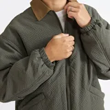 Campera de estilo urbano color verde oliva con textura acolchada, cierre frontal completo, cuello camisero en color marrón y puños elásticos.