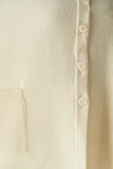 Capa color crudo de tejido de punto 100% lana fina, con cuello alto, cierre frontal con botones, bolsillos delanteros y corte asimétrico.