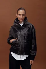 Chaqueta de cuero estilo bomber en color negro, con cuello alto, cierre frontal con cremallera y solapa con botones a presión. Presenta puños y cintura de tejido elástico acanalado, bolsillos laterales y un diseño estructurado.