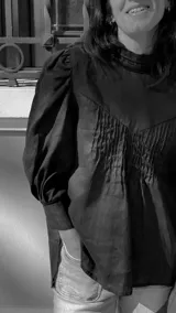 Blusa de lino color salmón con cuello alto, mangas abullonadas y detalle de alforzas en el frente. Presenta terminaciones con puntilla ondulada negra y cierre posterior con botones a la vista.