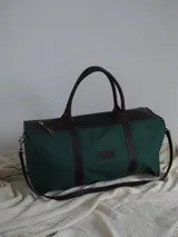Bolso tipo duffel de gran tamaño, confeccionado en lona color verde oliva con detalles y correas en cuero color suela. Cuenta con cierre superior y etiqueta de cuero con el logo de la marca.