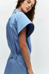 Vestido corto de estilo camisero color azul, con cuello solapa, manga corta y cierre frontal con botones. Presenta un bolsillo de plastrón con solapa en el pecho y un cinturón ajustable del mismo tejido en la cintura.