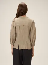 Camisa de lino mezcla color beige, con cuello redondo, cierre frontal con botones y mangas tres cuartos con puños elásticos. Presenta detalles de pespuntes verticales en zigzag en el frente.