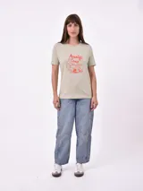 Remera de manga corta color beige claro con cuello redondo. Presenta un estampado gráfico central con un plato de pasta rodeado por un borde azul y blanco, sobre un fondo de franjas verticales rojas y blancas. El estampado incluye texto en cursiva en tonos turquesa y rojo.